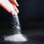 Poudre blanche granuleuse versée d'une main sur surface sombre